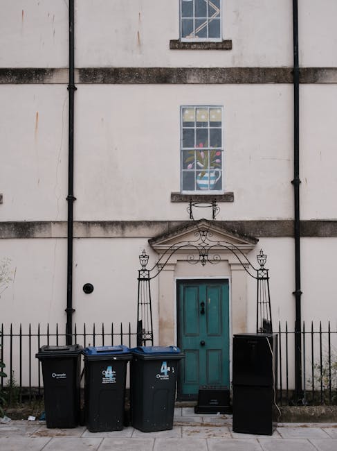 The image shows the facade of a white, slightly weathered building with a central turquoise door topped by a decorative wrought iron arch. Above the door, there is a large window with a smaller window