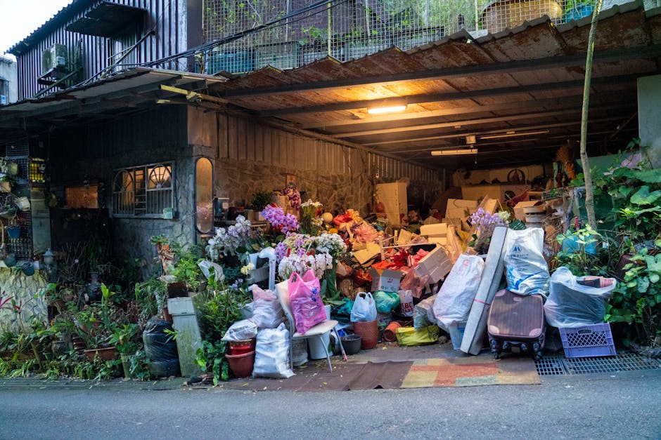 A cluttered outdoor area under a metal roof, filled with numerous flower arrangements and bouquets, including pink, purple, and white blossoms, some in vases and others wrapped in paper or plastic. Th