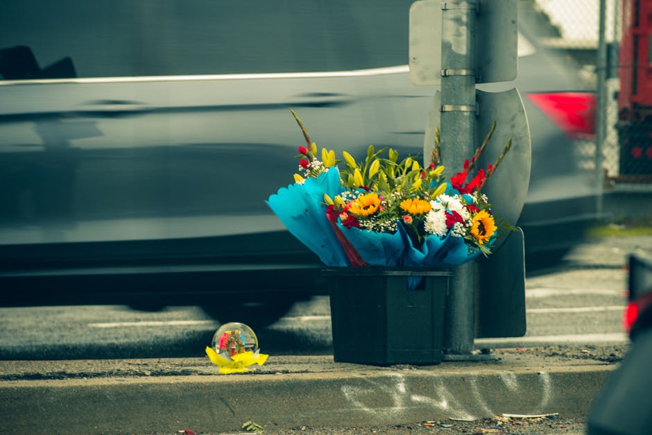 A colorful floral arrangement in a black plastic bucket, featuring yellow sunflowers, red and white blooms, and green foliage, wrapped in light blue paper with a red ribbon. The bouquet is placed on t