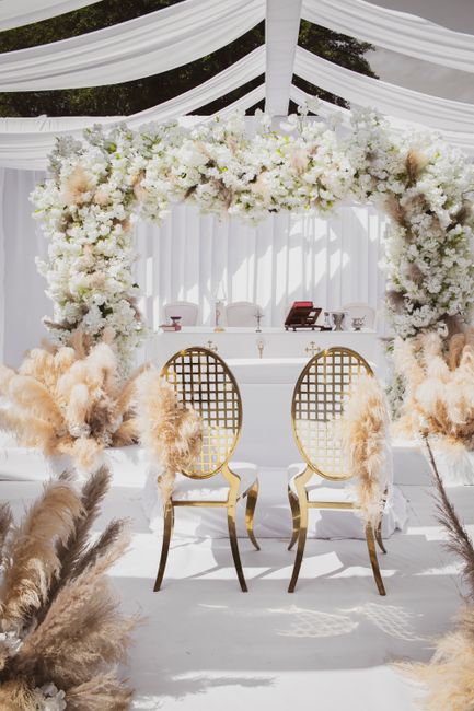 A beautifully arranged floral arch featuring predominantly white flowers, including roses, peonies, and hydrangeas, complemented by soft blush and cream accents. The arch is densely decorated with lus