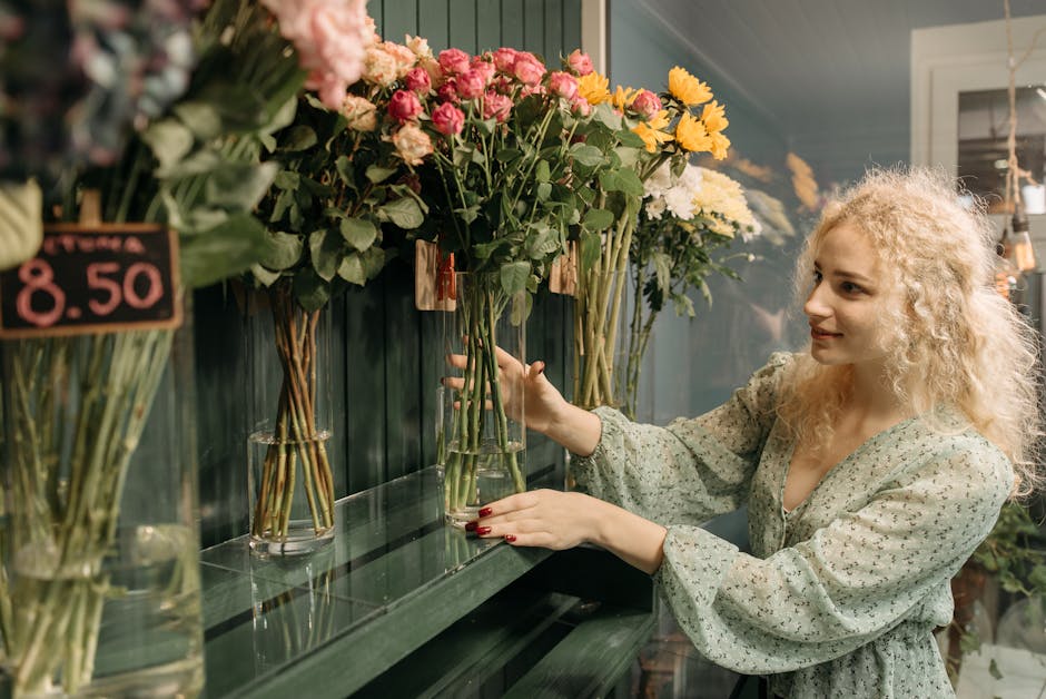 A young woman with curly blonde hair and fair skin is selecting a floral gift at a flower shop. She is wearing a light-colored, patterned blouse with puffy sleeves and is positioned to the right of th