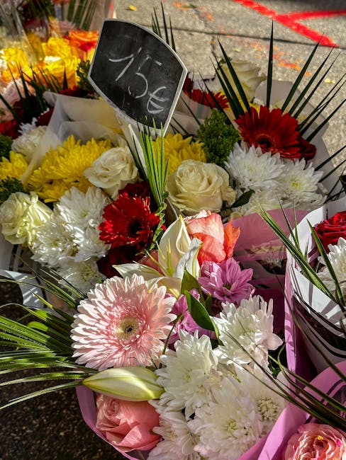 A vibrant floral arrangement featuring a mix of fresh flowers including large white roses, pink gerbera daisies, red gerbera daisies, and white carnations, with green foliage accents. The bouquet is w