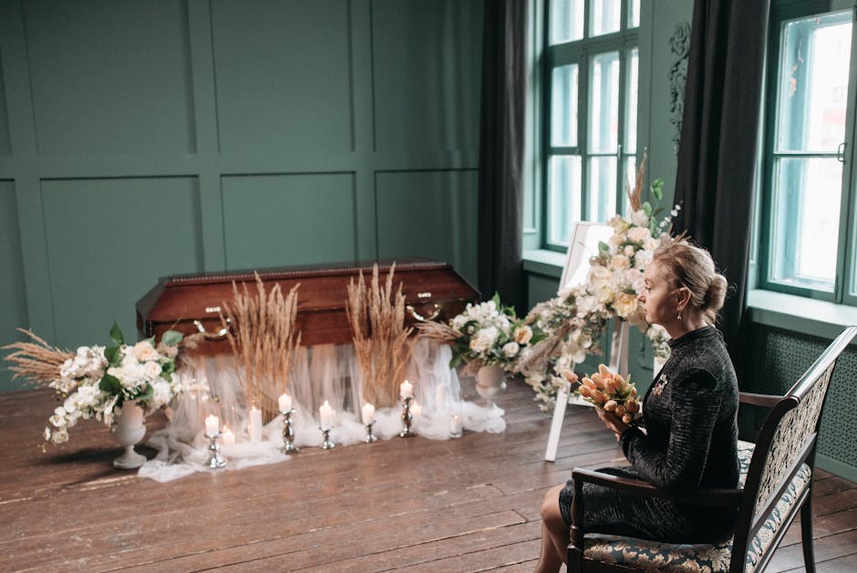 A woman seated in a floral arrangement setting indoors, holding a bouquet of soft peach and cream flowers, with her hair styled in an elegant updo. In front of her, there is a large arrangement of whi
