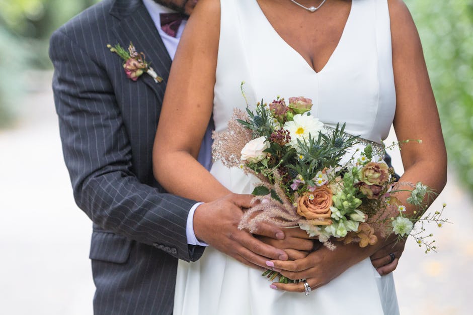 Close-up of a bride and groom holding a floral bouquet at an outdoor wedding. The bouquet features a mix of peach, white, and purple flowers, including roses and dainty daisies, complemented by green