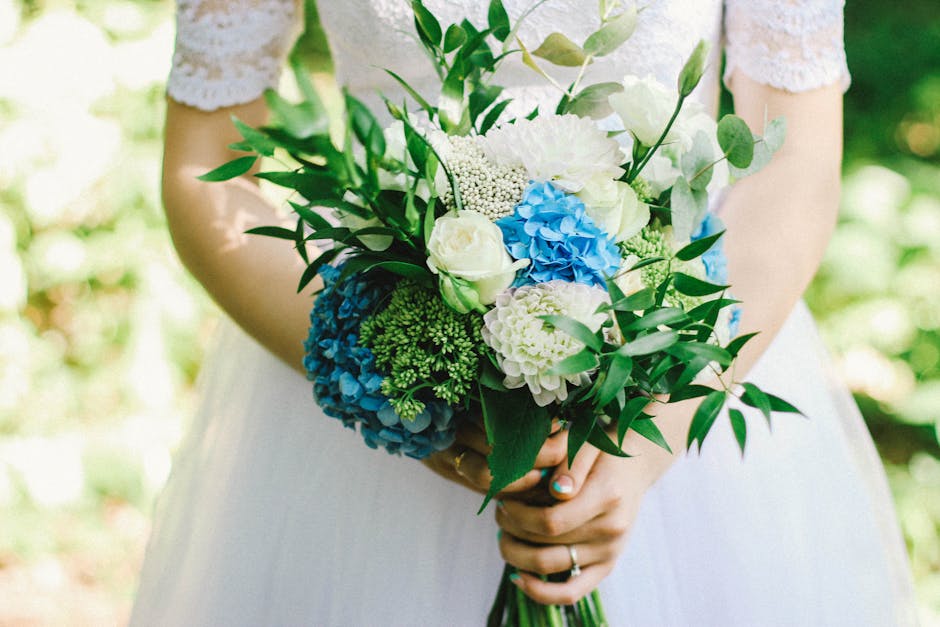 A bride holds a fresh floral bouquet featuring white roses, white peonies, and vivid blue hydrangeas, accented with green foliage including eucalyptus leaves. The bouquet is arranged in a loose, round